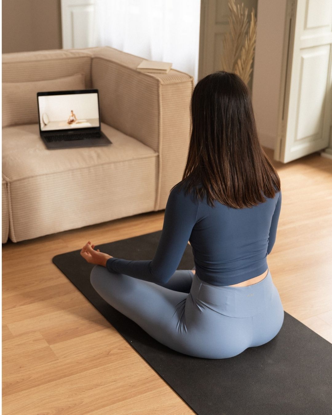 Mujer meditando sobre su esterilla en casa mientras sigue una clase de yoga online en el portátil